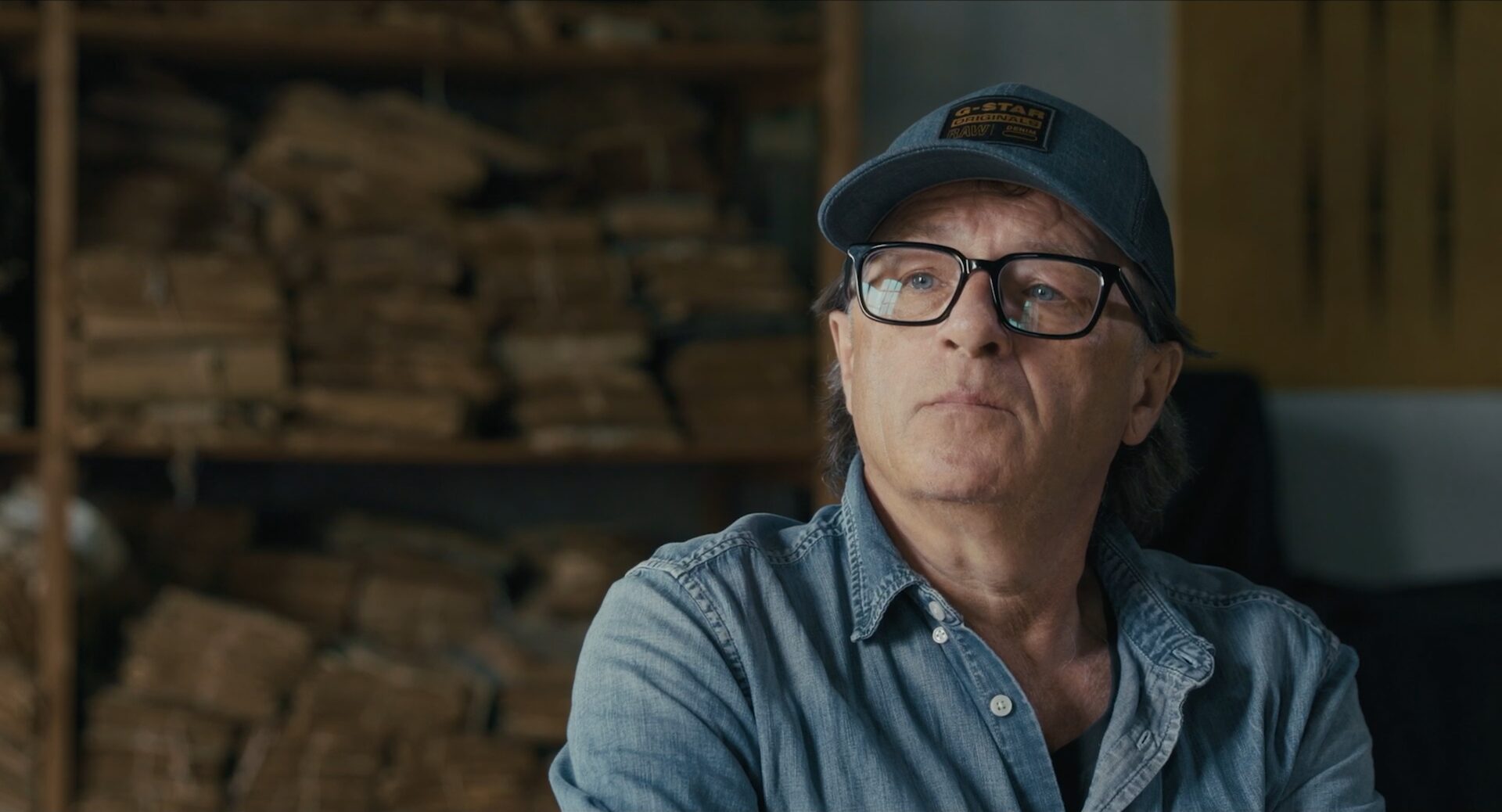 Aram Radomski mit Schirmmütze und Brille vor einem Regal voller Papierstapel