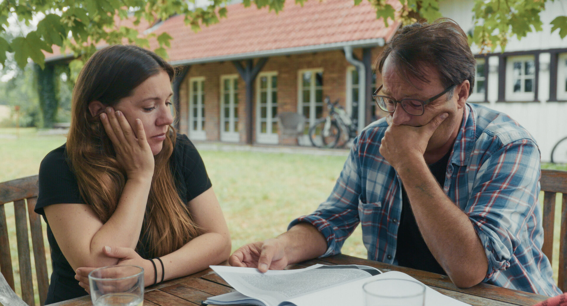 Aram Radomski und eine junge Frau sitzen nachdenklich an einem Gartentisch über Unterlagen